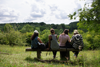 Four women sitting on a bench in a meadow looking out towards some trees