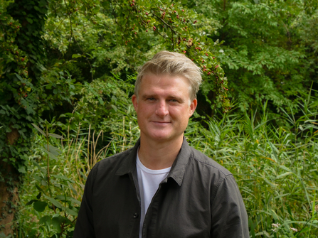 A man standing in front of a green backdrop full of vegetation. The person pictured is David Mooney