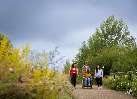 Four people making their way down a path surrounded by vegetation, the person in the middle pushes someone in a wheelchair and the two others walk either side